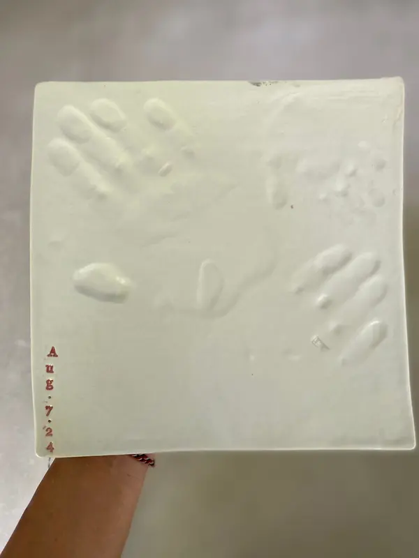 White square ceramic tile with two embossed handprints and the date Aug 7, 2024 stamped in red.