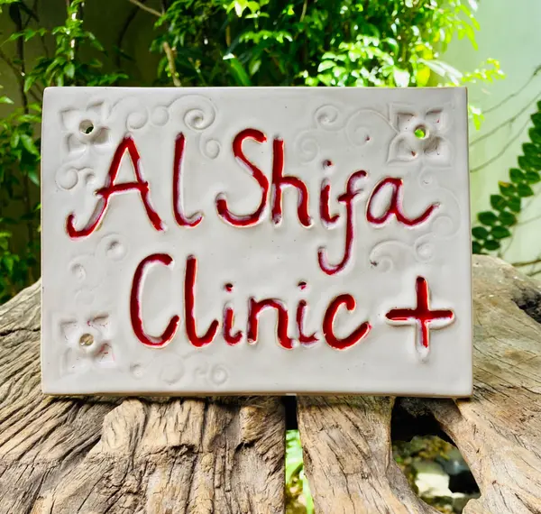 Rectangular white ceramic sign with red lettering reading 'Al Shifa Clinic +' and embossed scroll details.