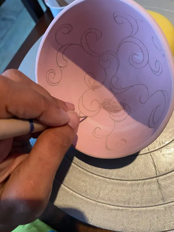 Hand carving scroll patterns into the interior of a pink ceramic bowl with a fine metal tool.