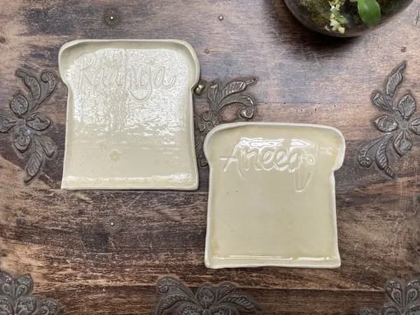 Two toast-shaped ceramic plates with names Raahya and Aneeq, in a glossy cream glaze on a dark wooden surface.