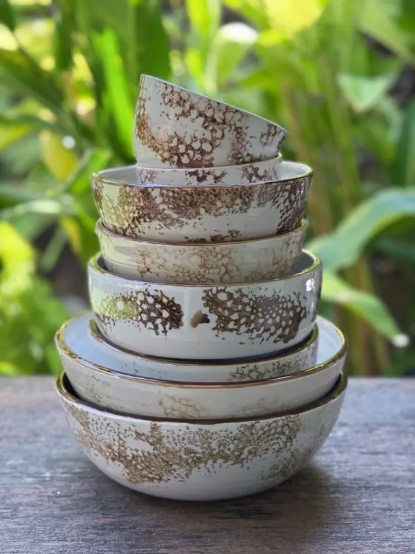 Stack of nested white ceramic bowls with a textured brown speckled glaze on a wooden surface against greenery.