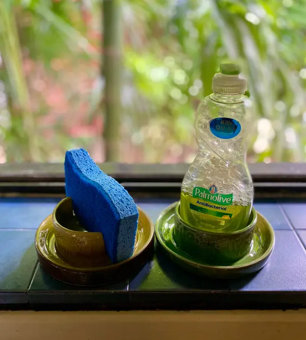 Green glazed ceramic sponge holder and soap bottle stand on blue tiled windowsill.