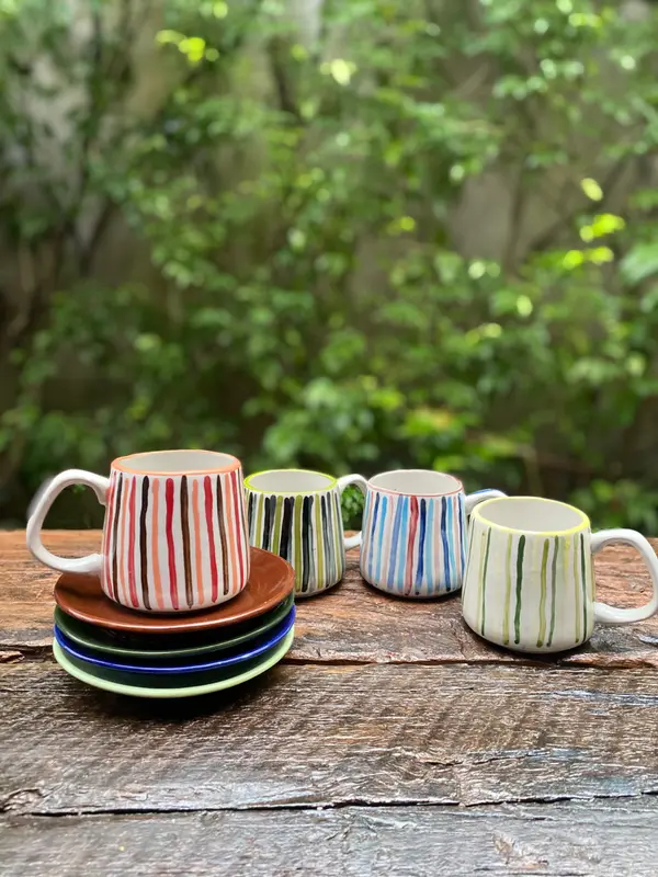 Four white ceramic mugs with colorful hand-painted vertical stripes on a wooden table with matching saucers.