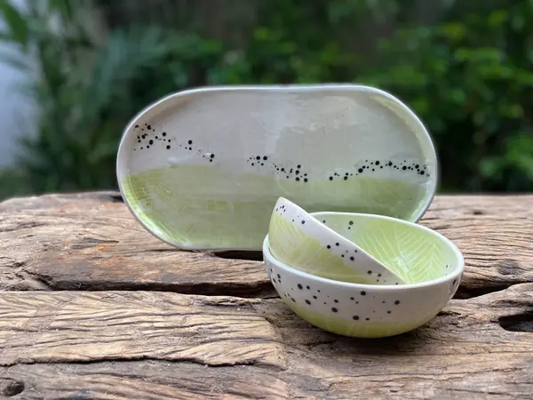 Handmade ceramic platter and bowls featuring light green leaf patterns and black speckles on a wooden surface.