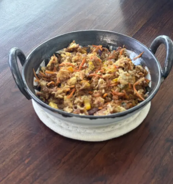 Black and white ceramic thaachiya bowl with twin handles, containing a stir-fry on a wooden table.