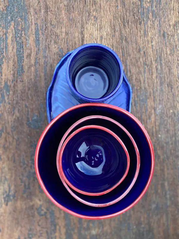 Top-down view of three nested cobalt blue bowls with red rims and a matching mug on a wooden background.