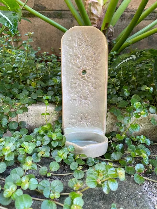 Sage green ceramic wall font with floral textures and a small basin, displayed in a garden setting.