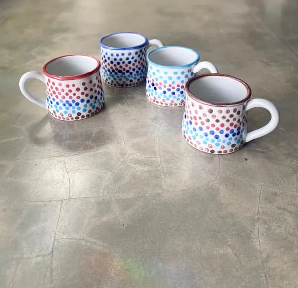 Four white ceramic mugs with colorful hand-painted polka dots and colored rims on a grey background.