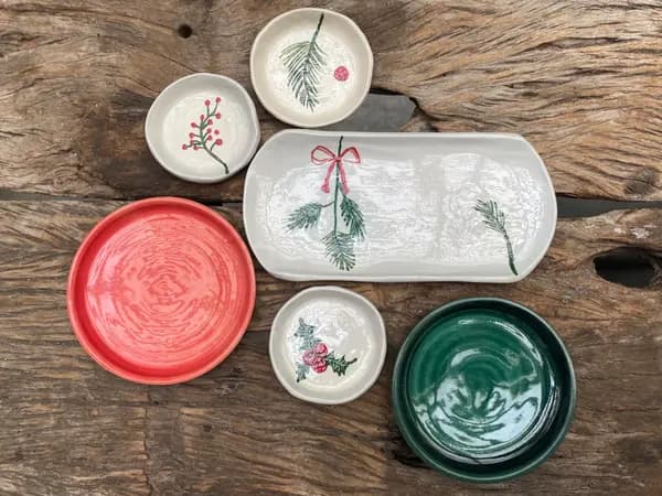 Assorted ceramic plates and platters with red, dark green, and winter botanical designs on a rustic wood surface.