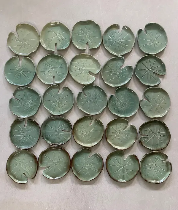 Grid of 25 sage green ceramic lily pad dishes with textured leaf veins on a neutral surface.