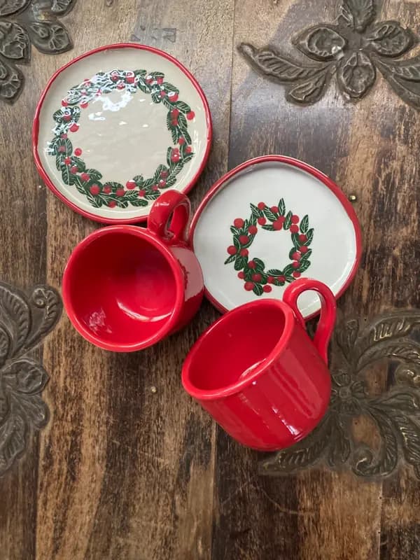 Two vibrant red ceramic mugs paired with white plates featuring painted berry wreaths on a wooden table.