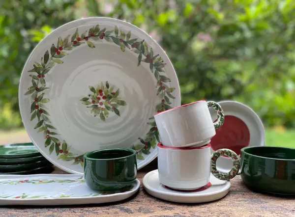 Hand-painted white ceramic dinnerware set featuring green leaf wreaths and red berries on a wooden surface.
