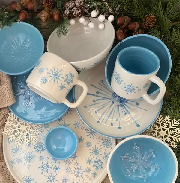 Ceramic mugs, bowls, and plates featuring light blue and white glazes with snowflake patterns and winter foliage.