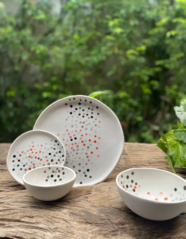 White ceramic plates and bowls with colorful hand-painted dots on a wooden table with a green foliage background.