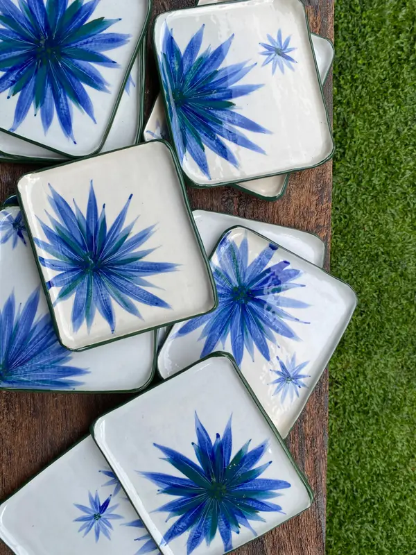 Square white ceramic plates featuring blue starburst floral designs and dark green rims on a wooden table.