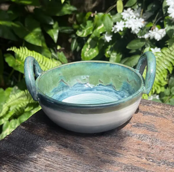 Two-handled ceramic bowl with layered turquoise and green glazes over a white base, placed on a wooden surface.