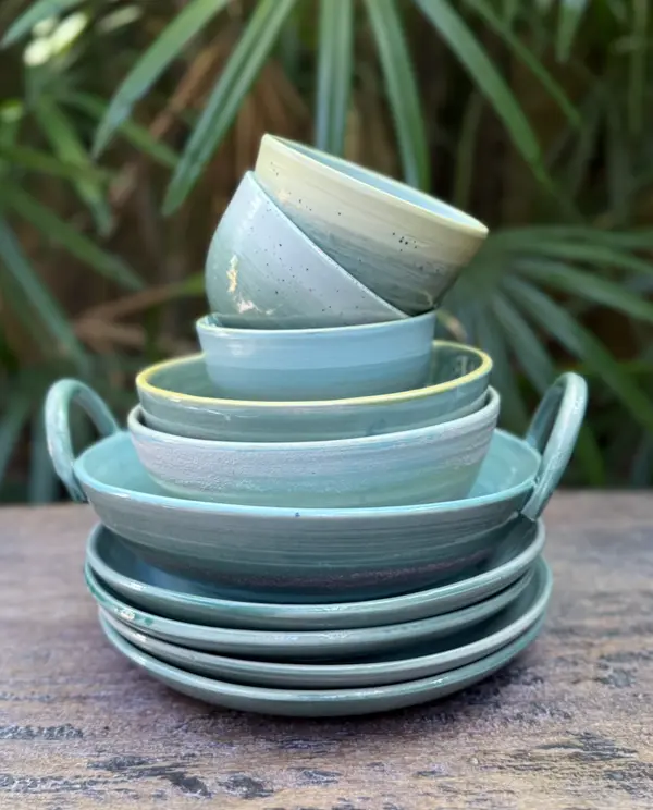 Stack of light green and turquoise ceramic bowls and plates on a wooden surface with a tropical leaf background.