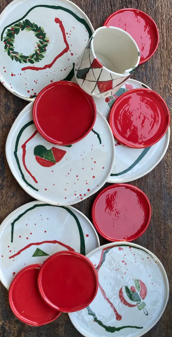 Hand-painted white ceramic plates and mug with festive red and green designs, including a wreath and splatter details.