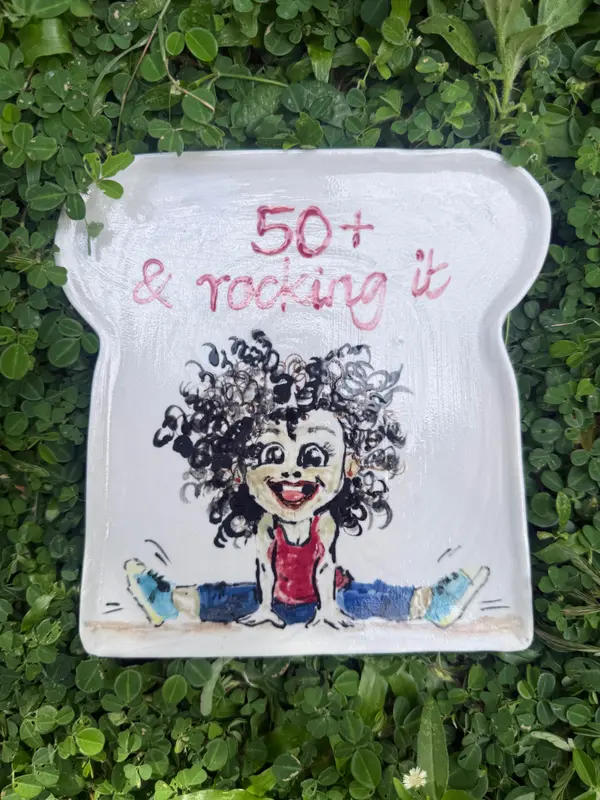 Toast-shaped white ceramic plate with a hand-painted cartoon woman and pink text reading 50+ & rocking it.