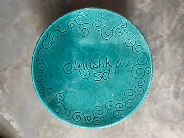 Turquoise crackle-glaze ceramic disc with engraved scrolls and text reading 'Anushka 50th' in the center.