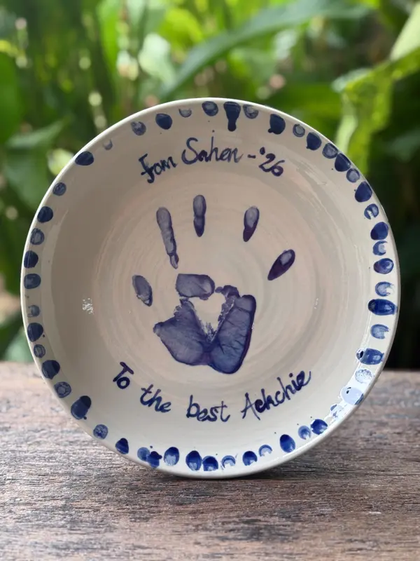 White ceramic plate with a cobalt blue handprint, dotted border, and handwritten blue text on a wooden surface.