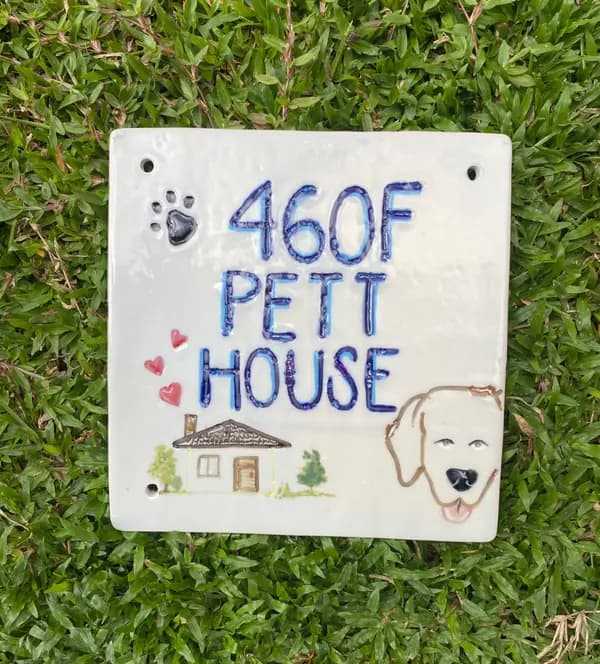 A white square ceramic house number tile featuring a dog portrait, blue text, and small house illustration.