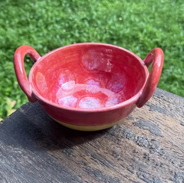Red ceramic bowl with two side handles and a vibrant pink-mottled interior glaze on a weathered wooden surface.