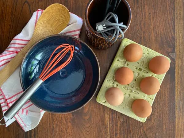 Dark blue glazed bowl, textured brown utensil holder, and light green egg tray styled with kitchen tools on wood.