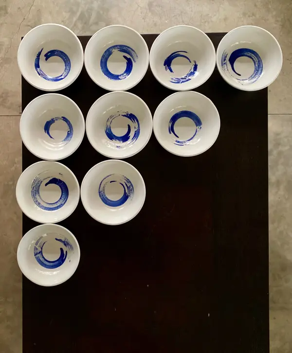 Ten white ceramic bowls with hand-painted cobalt blue enso circles, arranged on a dark surface.