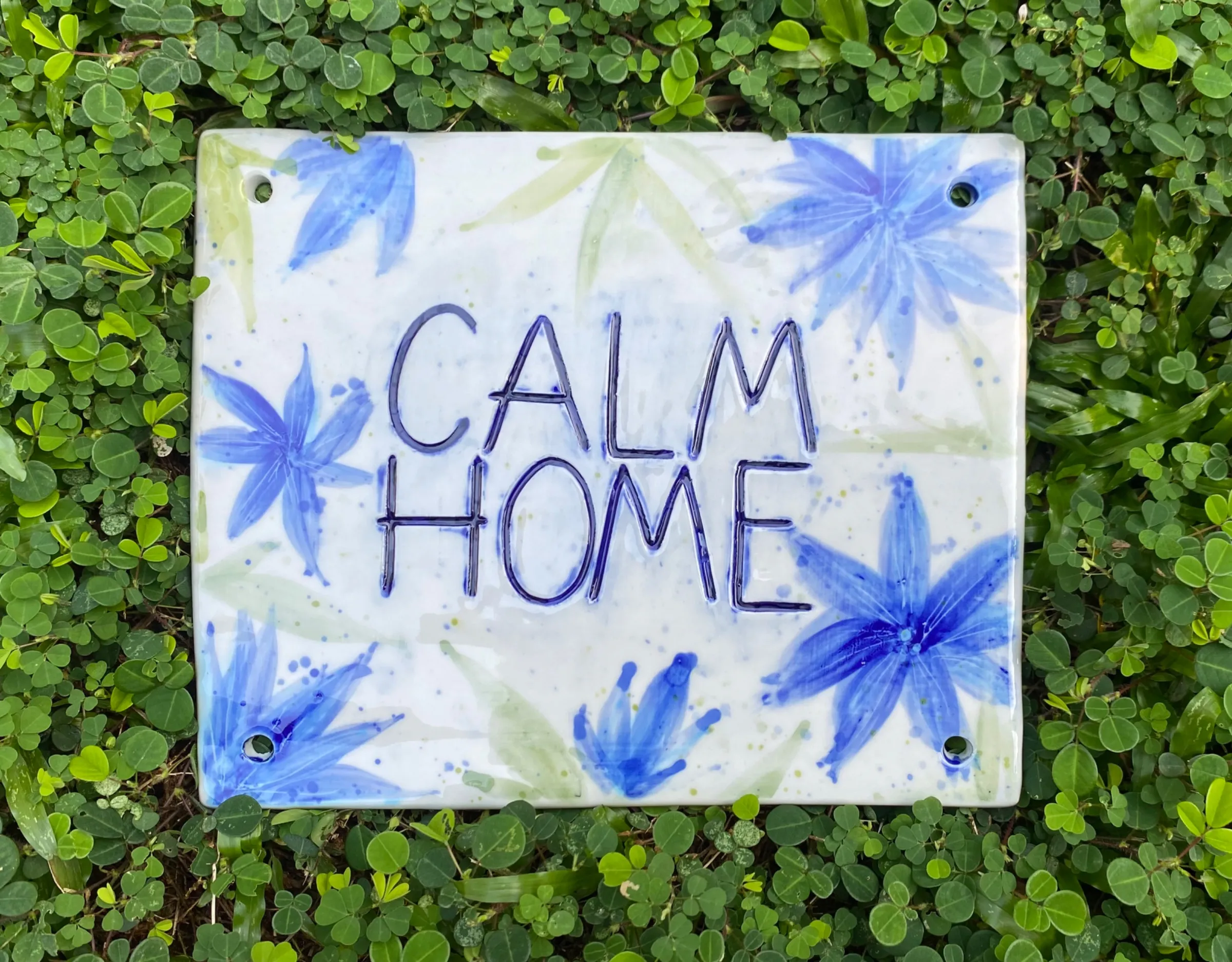 Square ceramic plaque with 'CALM HOME' text and blue floral designs, resting on green leaves.