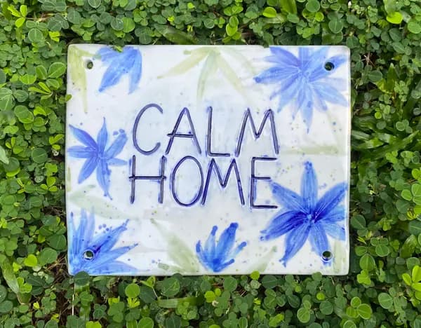 Square ceramic plaque with 'CALM HOME' text and blue floral designs, resting on green leaves.