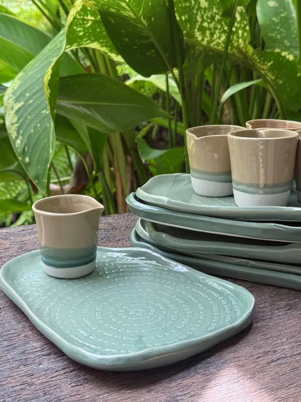 Handmade sage green textured plates and small tri-color glazed jugs on a wooden table with lush greenery behind.