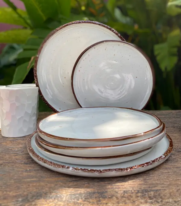 Handmade speckled white plates with brown rims stacked beside a faceted white cup on a rustic wood surface.