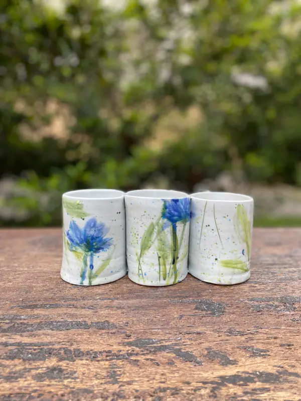 Three white ceramic tumblers with blue floral watercolor-style painting on a rustic wooden surface.