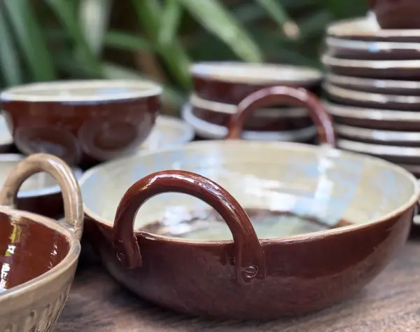 Brown glazed ceramic thaachiya bowl with loop handles among other stacked bowls on a wooden surface.