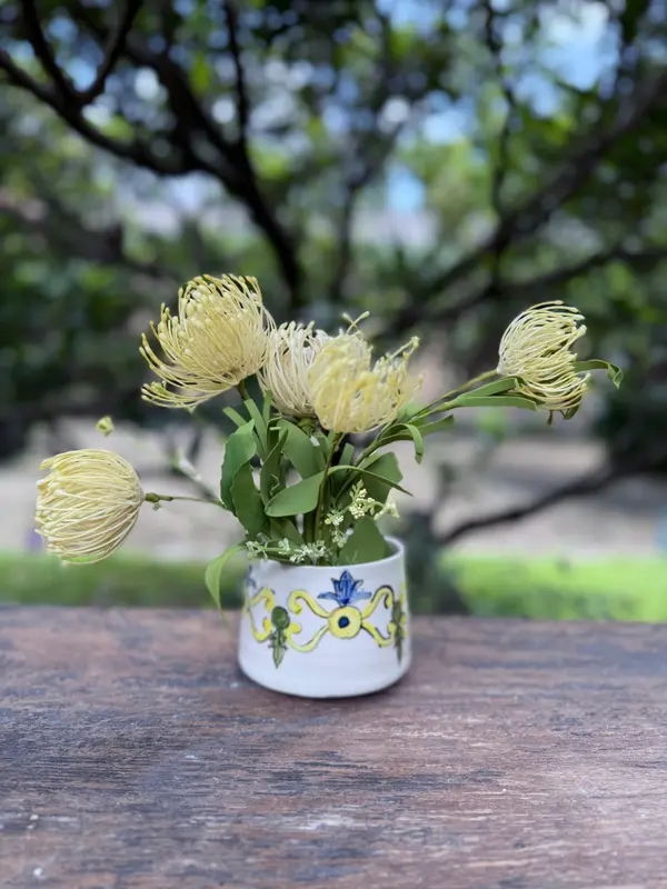 Small white ceramic vase with yellow and blue hand-painted patterns holding yellow pincushion flowers on wood.