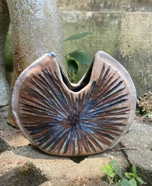 Textured ceramic disk vase with radiating brown and blue grooves shaped like a water lily leaf on stone.