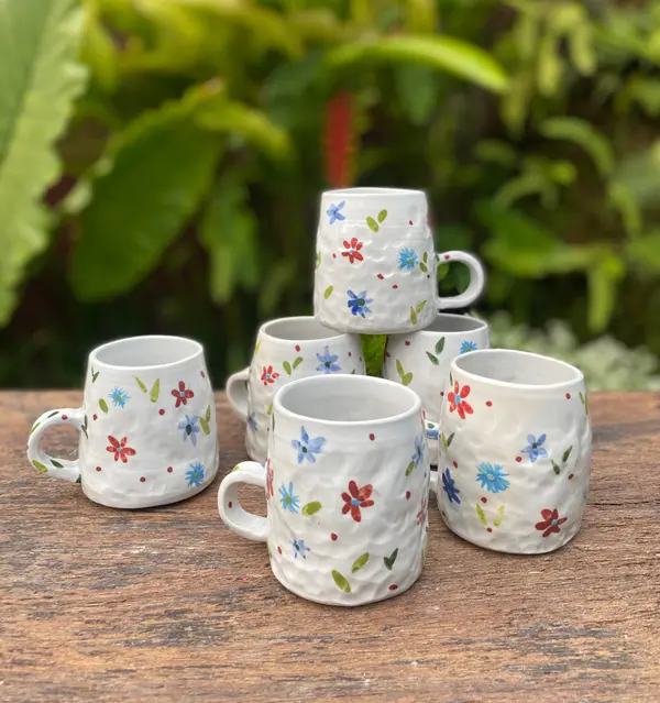 Six white textured ceramic mugs with hand-painted red and blue flowers on a wooden table outdoors.