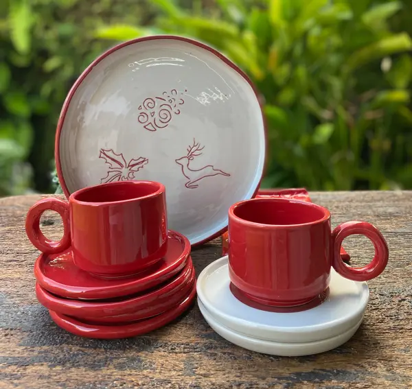 Red mugs and saucers with a white Christmas-themed reindeer plate on a wooden surface outdoors.