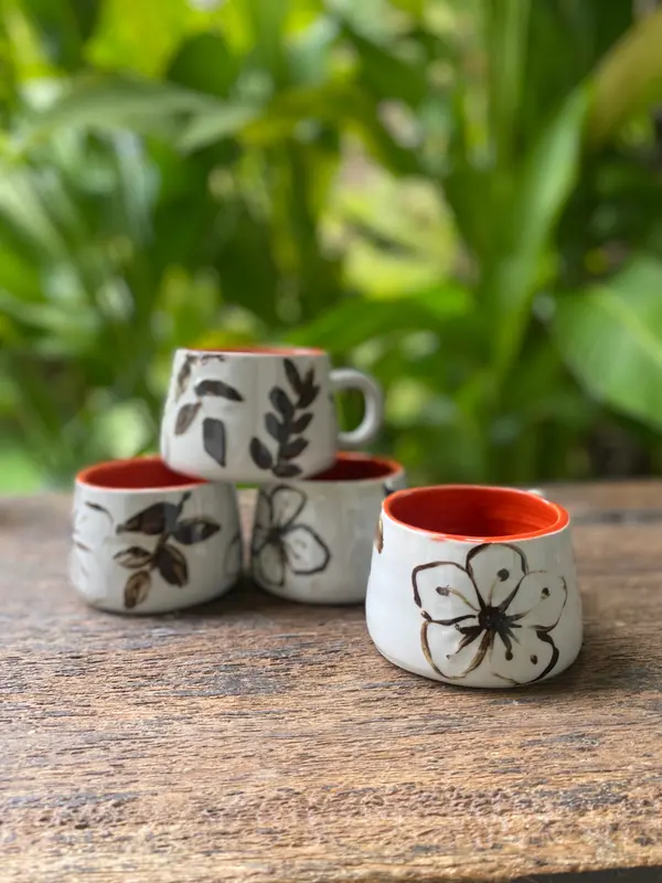 Four white mugs with black botanical sketches and bright orange interiors on a wooden surface.