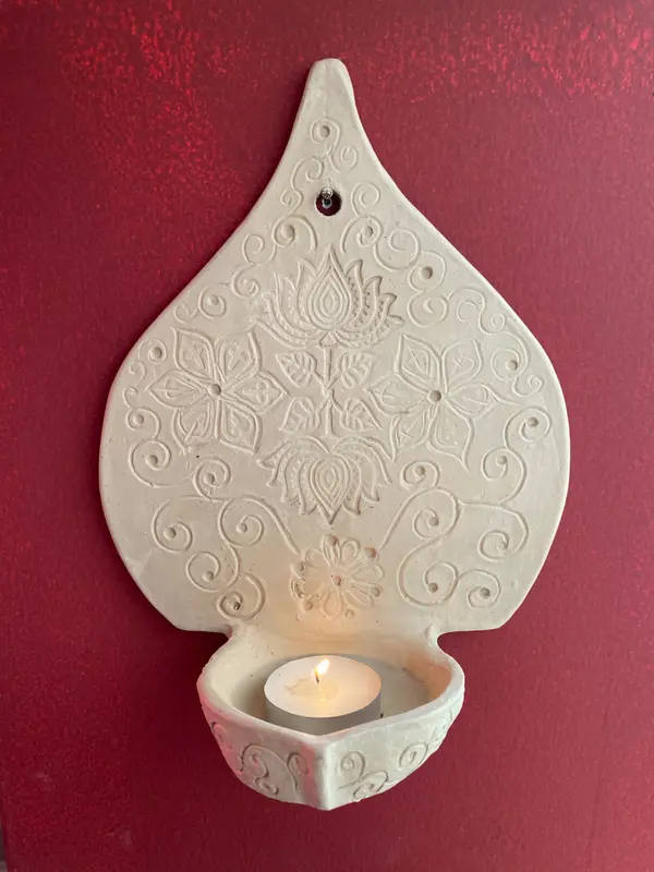 Beige ceramic wall-mounted candle holder with carved floral patterns and a lit tealight on a red background.