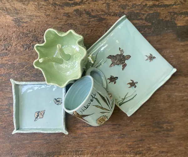 Assorted light green and blue pottery including a turtle platter, shell plate, and mug with ocean motifs on wood.