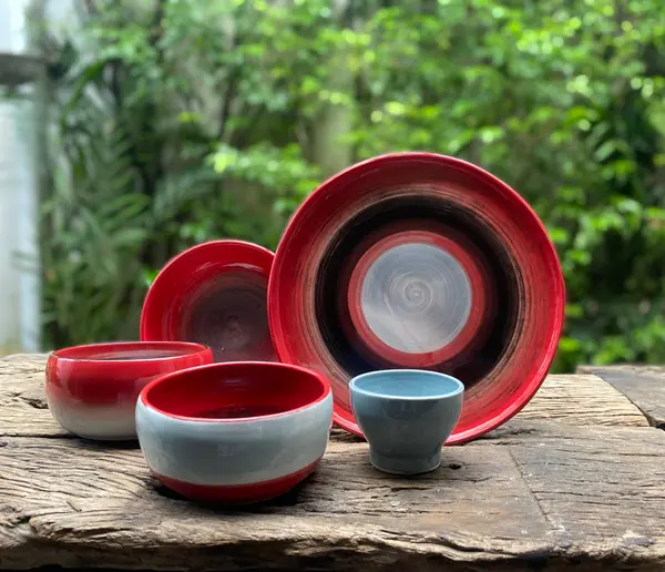 Ceramic pottery set with red, black, and light blue concentric glazes on a rustic wooden surface against greenery.