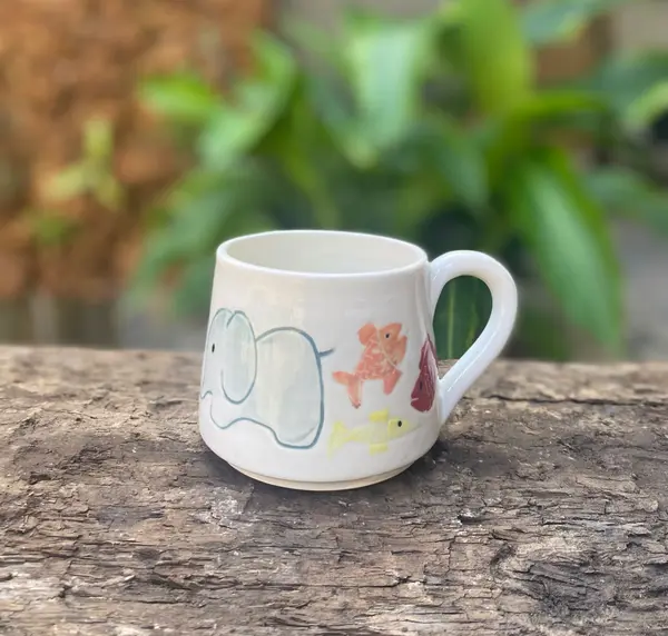 White ceramic mug featuring hand-painted light blue elephant and colorful fish on a rustic wood background.