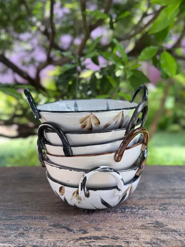 Stacked white ceramic bowls with side handles and painted leaf patterns, set on a wooden table.