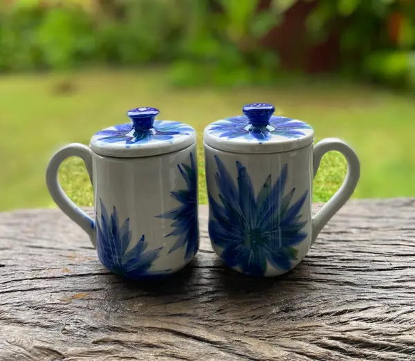 Two white ceramic mugs with lids featuring hand-painted cobalt blue floral designs on a rustic wooden table.