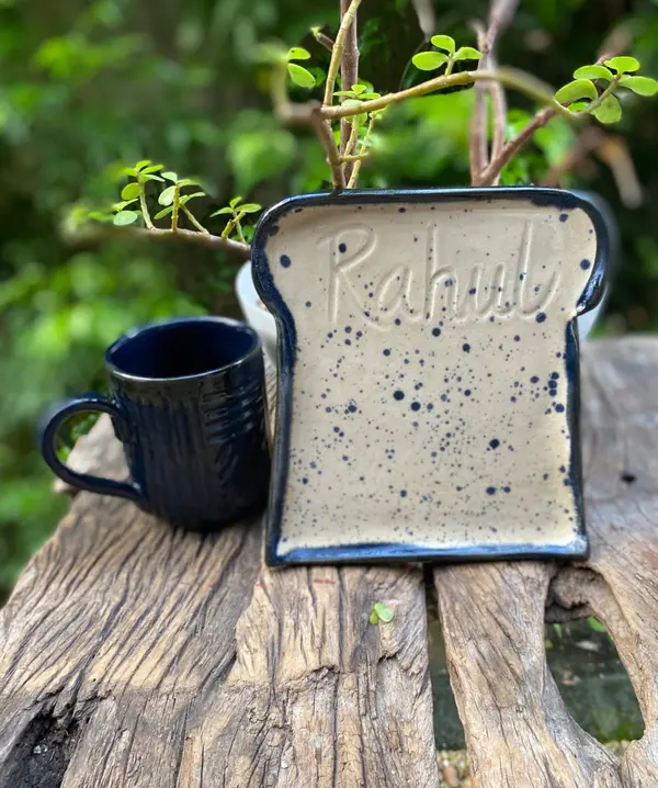 A toast-shaped ceramic plate with the name Rahul, speckled beige glaze, and a dark blue mug on weathered wood.