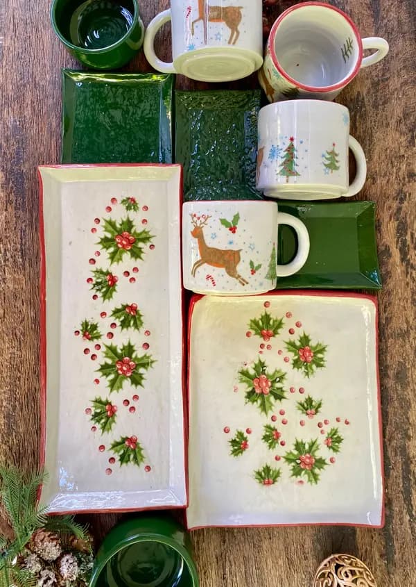 Holiday pottery set featuring mugs and platters with hand-painted reindeer and holly designs on white and green glaze.