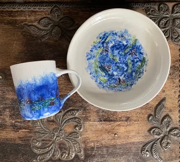 Hand-painted ceramic mug and bowl set with blue water lily designs on a carved wooden surface.