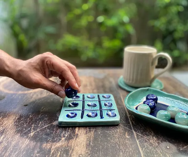 Ceramic tic-tac-toe set with turquoise board and cobalt blue and green pieces on a wooden table.